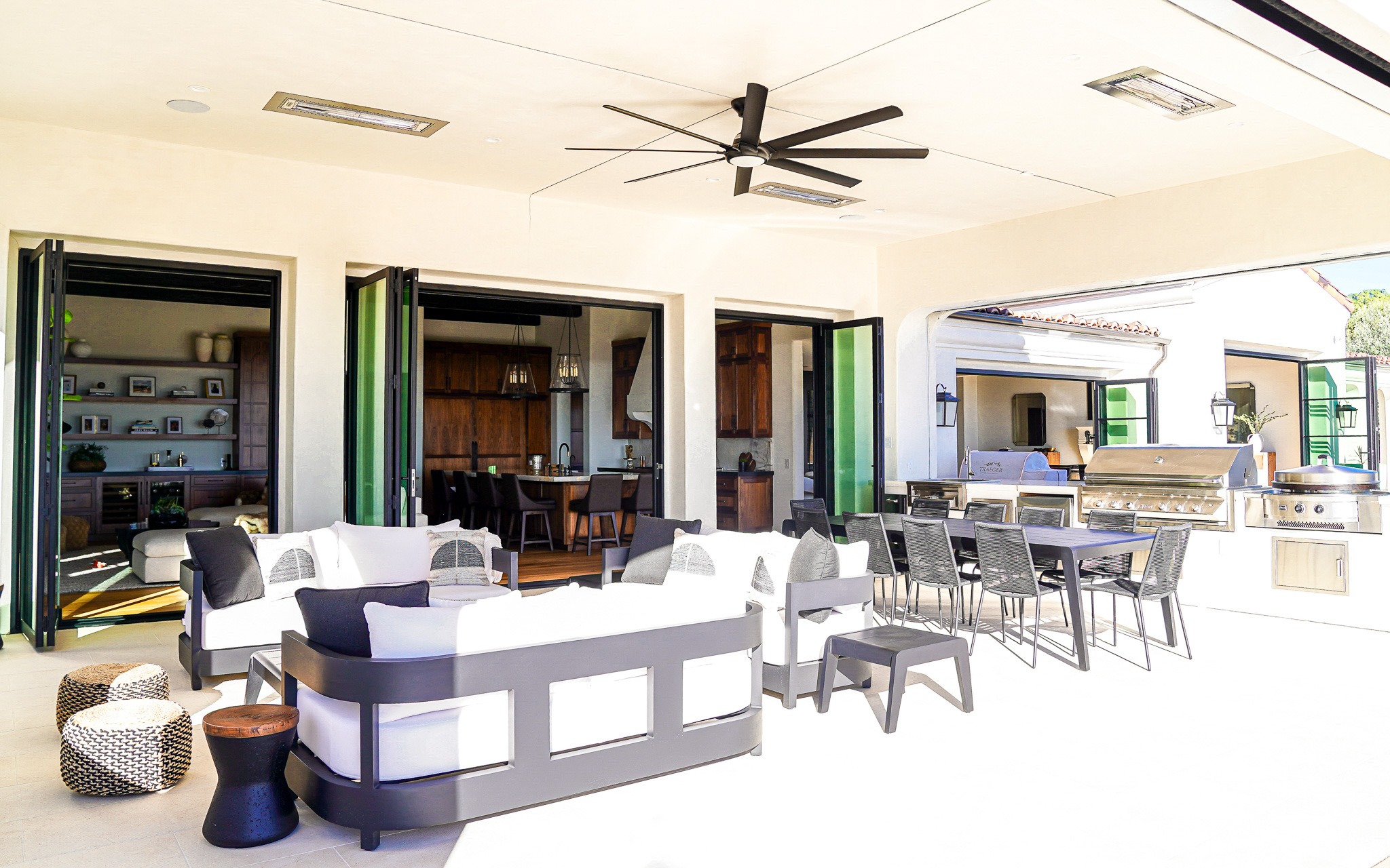 Untitled-233 Luxurious outdoor patio area with a modern seating arrangement, ceiling fans, open folding doors, and an integrated kitchen with bar stools.