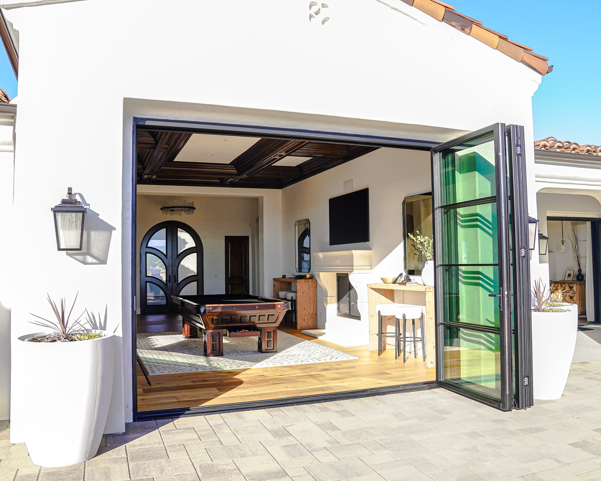 Untitled-237 Modern home patio with large sliding glass doors open to a stylish interior living space, featuring a kitchen island, wood-beamed ceiling, and furnishings.