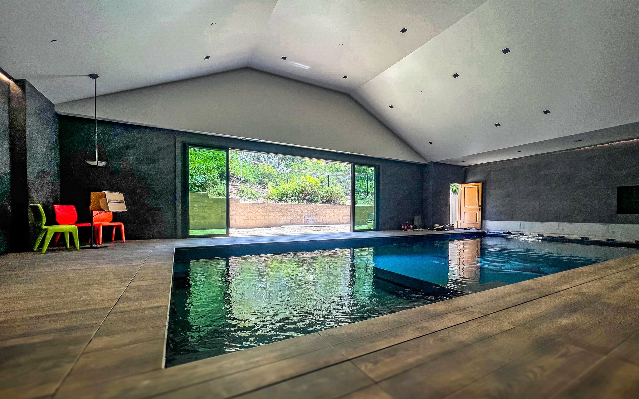 Untitled-65 Modern indoor swimming pool with dark gray walls and large windows overlooking a garden. there's a colorful seating area near the windows.