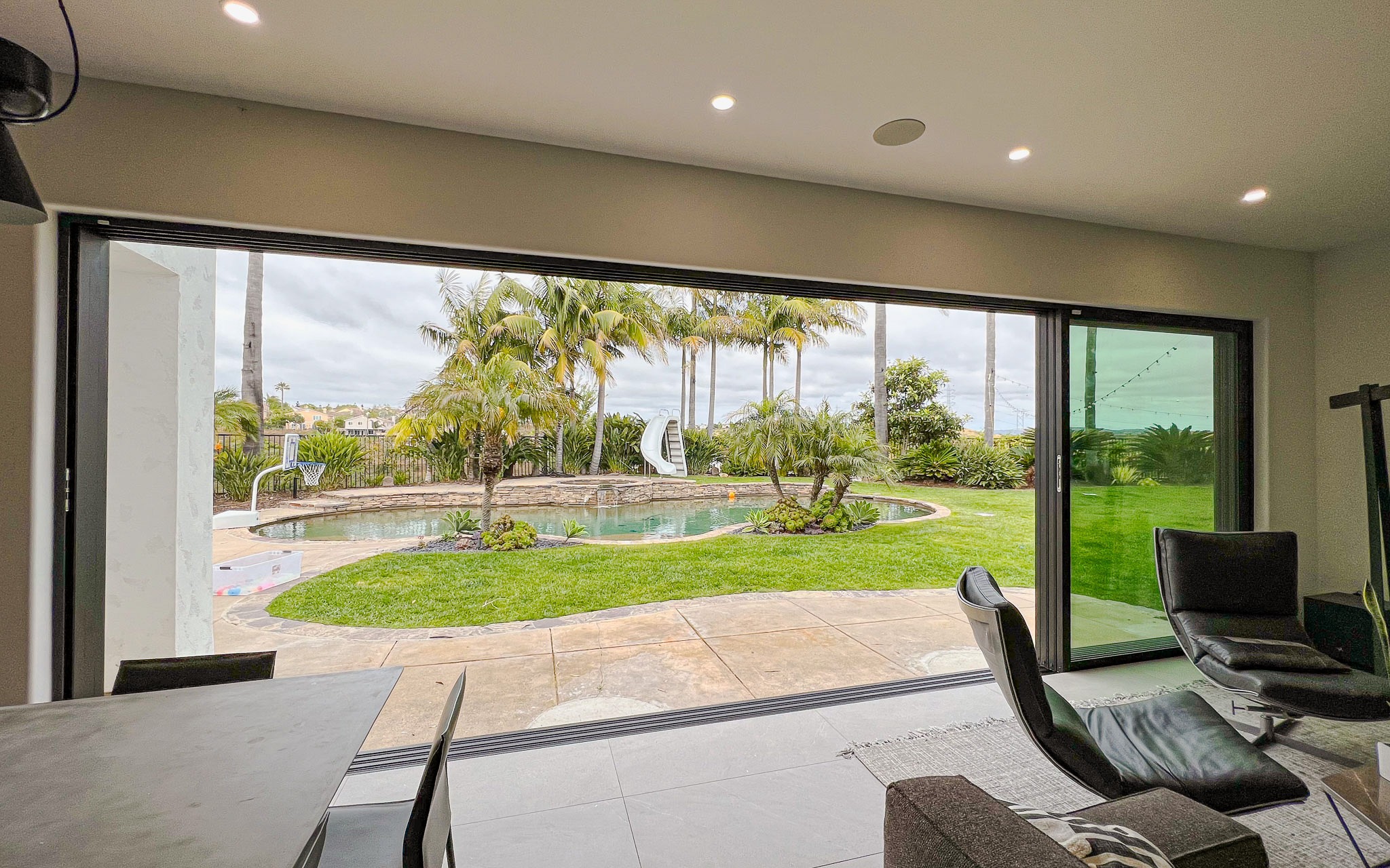 Untitled-78 Modern living room with large sliding glass door overlooking a backyard with a swimming pool and tropical landscaping.